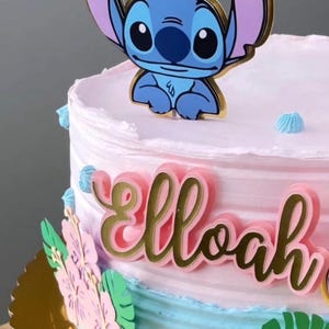 Puede incluir: Una tarta decorada en rosa y azul, con un adorno de personaje de dibujos animados. El nombre "Elloah" se muestra en letras doradas. Decoraciones florales en rosa y verde adornan el lateral de la tarta.