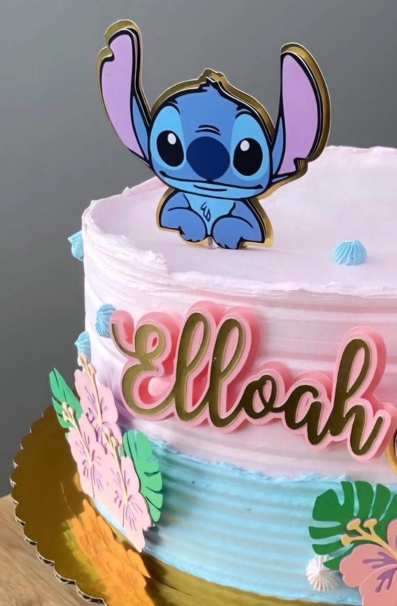Puede incluir: Una tarta decorada en rosa y azul, con un adorno de personaje de dibujos animados. El nombre "Elloah" se muestra en letras doradas. Decoraciones florales en rosa y verde adornan el lateral de la tarta.