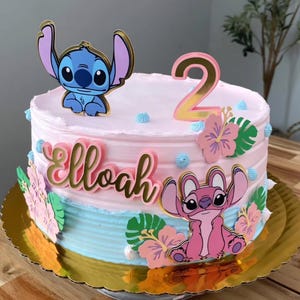 Puede incluir: Tarta de dos pisos con glaseado rosa y azul, decorada con personajes de dibujos animados, flores y el n&uacute;mero 2. El nombre "Elloah" est&aacute; escrito en letras doradas. La tarta est&aacute; sobre una base dorada y festoneada.