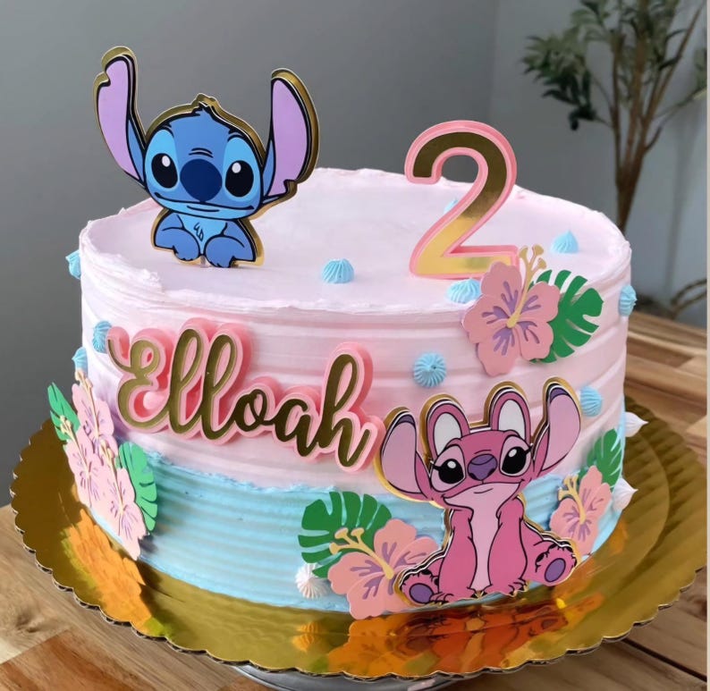 Puede incluir: Tarta de dos pisos con glaseado rosa y azul, decorada con personajes de dibujos animados, flores y el n&uacute;mero 2. El nombre "Elloah" est&aacute; escrito en letras doradas. La tarta est&aacute; sobre una base dorada y festoneada.