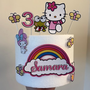Puede incluir: Una tarta blanca decorada con Hello Kitty, Keroppi, My Melody y recortes de mariposas. La tarta está adornada con un arcoíris y el nombre "Samara". Un gran número rosa "3" indica la edad del cumpleaños.