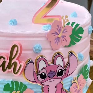 Puede incluir: Un pastel glaseado rosa decorado con un adorno para pastel con el n&uacute;mero 2 dorado y rosa. El pastel est&aacute; adornado con un personaje de dibujos animados rosa, flores rosas y hojas verdes. Los acentos de glaseado azul a&ntilde;aden un toque de color.