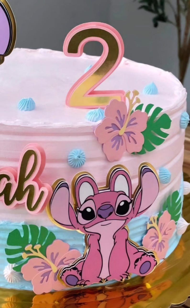 Puede incluir: Un pastel glaseado rosa decorado con un adorno para pastel con el n&uacute;mero 2 dorado y rosa. El pastel est&aacute; adornado con un personaje de dibujos animados rosa, flores rosas y hojas verdes. Los acentos de glaseado azul a&ntilde;aden un toque de color.