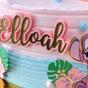 Puede incluir: Un pastel decorado con glaseado rosa y azul, con una placa con el nombre rosa con borde dorado que dice "Elloah". El pastel est&aacute; adornado con recortes de personajes de dibujos animados y decoraciones florales.
