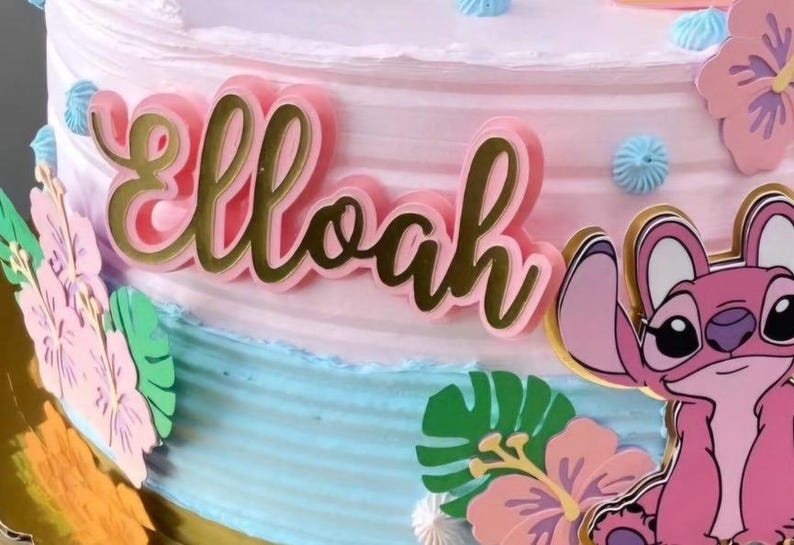 Puede incluir: Un pastel decorado con glaseado rosa y azul, con una placa con el nombre rosa con borde dorado que dice "Elloah". El pastel est&aacute; adornado con recortes de personajes de dibujos animados y decoraciones florales.