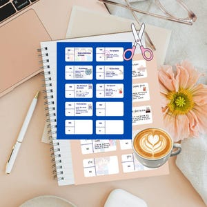 May include: A spiral-bound notebook with a blue and white grid of text boxes, a pen, and a cup of coffee with latte art. A pair of pink scissors and a flower are also present.