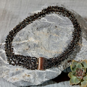 Może przedstawiać: Wielożyłowy naszyjnik z koralików w odcieniach brązu i czerni, z zapięciem w kolorze miedzi. Naszyjnik jest prezentowany na szarej skale, z rośliną sukulentową w tle. Koraliki wydają się małe i blisko siebie.