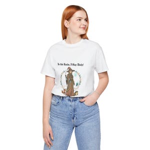 Op de afbeelding: Wit T-shirt met een illustratie van een heks in een bruine jurk, omringd door een vos en een egel. De tekst "In this Realm, I Slay Daily!" staat erboven. Het shirt wordt gedragen met een blauwe jeans.