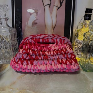 May include: A handmade, chunky-knit clutch in shades of red and pink. The bag features a rectangular shape with a small handle. The background includes a framed image of legs in high heels and decorative glass bottles.