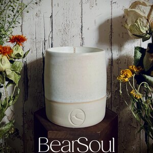 Puede incluir: Una vela de cerámica color crema en forma cilíndrica con un logotipo en la base. La vela está sobre un bloque de madera oscura, con flores secas y follaje en el fondo. El texto "BearSoul WELLBEING" está en la parte inferior.