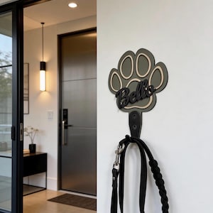 Könnte beinhalten: Ein Wandhaken in Form eines Pfotenabdrucks mit dem Namen Bello in schwarzer Schreibschrift. Die Pfote ist olivgrün mit einer hellbeigen Umrandung. Ein schwarzer Metallhaken ist unten befestigt und hält eine schwarze Leine mit silbernem Clip.