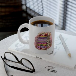 May include: White ceramic coffee mug with a stack of books illustration. The books are in various colors, with floral accents. The mug is filled with dark coffee. A pencil, glasses, and paperclips are also visible.
