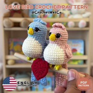 Könnte beinhalten: Zwei gehäkelte Liebesvögel, einer blau und einer rosa, sitzen auf einem Holzstab mit einem roten Herz. Die Vögel haben gelbe Schnäbel und Füße. Das Bild enthält den Text "LOVE BIRD CROCHET PATTERN" und "ENGLISH (PATTERN LANGUAGE)".