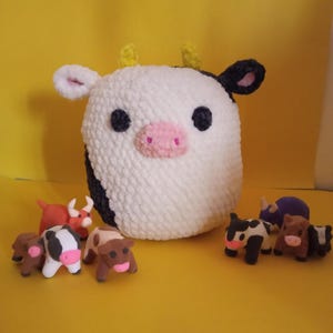 May include: A large, white, plush cow with black spots, black eyes, and a pink nose. Small, colorful cow figurines are arranged in front of the plush cow. The background is a solid yellow.