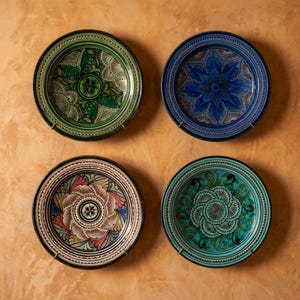 May include: A set of four round ceramic plates featuring intricate hand-painted patterns. The top left plate is green, the top right is blue, the bottom left is multi-colored with floral motifs, and the bottom right is teal with swirling designs.