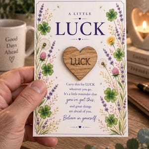 May include: A rectangular card featuring the text A LITTLE LUCK at the top and a wooden heart token engraved with the word LUCK in the centre. The card is decorated with illustrations of four-leaf clovers, lavender, and wildflowers.
