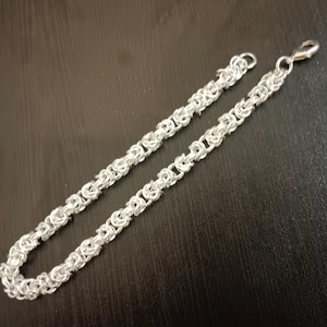 Puede incluir: Una pulsera de cadena de color plateado con un patrón de tejido bizantino construido a partir de anillos de metal entrelazados. La pulsera tiene un acabado metálico pulido e incluye un pequeño cierre de mosquetón en un extremo.