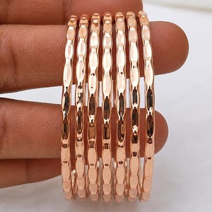 May include: A set of seven rose gold-toned metal bangles, each featuring a repeating vertical hammered texture along the outer surface. The bangles are thin, circular, and have a polished, reflective finish that catches the light evenly.