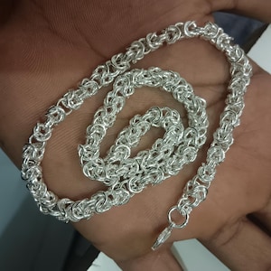 Op de afbeelding: Een glanzende zilveren ketting met een complex Byzantijns schakelpatroon. De metalen schakels zijn ingewikkeld met elkaar verweven om een gestructureerd, cilindrisch ontwerp te creëren dat licht reflecteert op het gepolijste oppervlak.