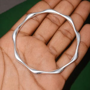 Puede incluir: Un brazalete de plata pulida que presenta un diseño ondulado y continuo. La superficie metálica es lisa y reflectante, formando una figura circular con curvas consistentes a lo largo de toda la circunferencia de la pieza de joyería.