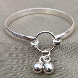 May include: A polished silver bangle bracelet featuring a circular clasp and two small, round dangling charms. The bracelet's design is simple and elegant. The charms are also silver, with a smooth, reflective surface. The bracelet is made of metal.