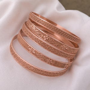 Pode incluir: Um conjunto de quatro pulseiras rígidas de metal em tom de ouro rosa, cada uma com padrões em relevo distintos, incluindo favos de mel geométricos, motivos florais, texturas pontilhadas e um design trançado contínuo, empilhadas para exibir.