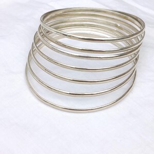 May include: A stack of seven thin, polished silver-coloured metal bangles. Each bangle features a smooth, rounded surface and a circular shape. The bracelets are arranged in a slightly angled, overlapping formation, creating a layered appearance.