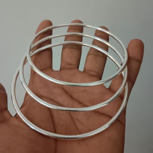 Puede incluir: Tres brazaletes circulares de tono plateado, lisos y pulidos, apilados juntos. Cada brazalete presenta un diseño de banda redonda simple y continua con un acabado metálico reflectante. El conjunto consta de tres anillos finos idénticos dispuestos en una formación en capas.