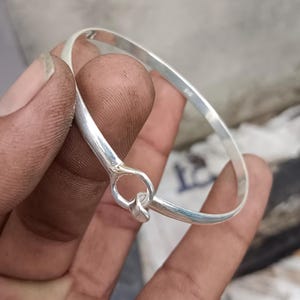 May include: A polished silver bangle bracelet featuring a hook and eye style closure mechanism. The metal band has a smooth, reflective surface and a circular loop detail where the two ends meet, with a small 925 hallmark stamped on the inner side.