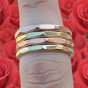 Könnte beinhalten: Ein Stapel von fünf Metallringen in verschiedenen Farben: Gold, Roségold, Silber und Hellgrün. Die Ringe befinden sich auf einem Finger, mit einem verschwommenen Hintergrund aus roten Rosen.