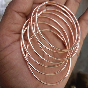 May include: A stack of multiple thin, circular bangles with a polished rose gold metallic finish. The bangles are smooth, round, and uniform in thickness, creating a layered appearance with reflective surfaces that catch the light.