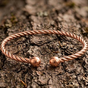 Peut inclure: Un bracelet manchette en métal de couleur cuivre présentant une texture torsadée semblable à une corde sur toute la longueur de l'anneau. Le bracelet est ouvert et se termine aux deux extrémités par une boule sphérique lisse et polie.