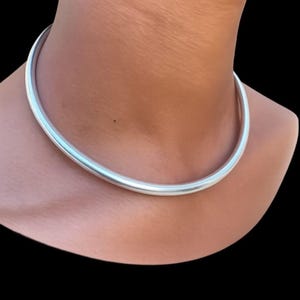 May include: A smooth, solid silver-toned metal choker necklace with a rounded, tubular profile. The necklace features a continuous, seamless design that sits flush against the neck, showcasing a polished, reflective surface with a clean finish.