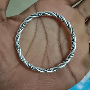 Può includere: Un bracciale rigido circolare in metallo color argento lucido, caratterizzato da un design a corda intrecciata continua. La superficie del metallo presenta qualità riflettenti ad alta lucentezza con accenti ossidati scuri nelle scanalature.