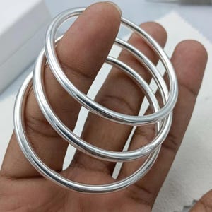 Peut inclure: Trois joncs en argent poli avec un profil lisse et arrondi. Chaque bracelet présente une finition métallique très brillante et un design circulaire simple. L'un des joncs porte clairement le poinçon 925, indiquant de l'argent sterling.
