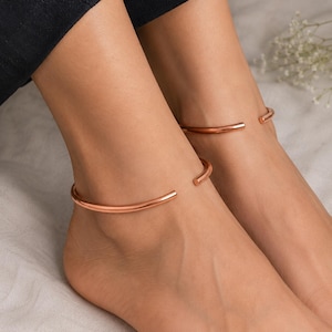 May include: A pair of polished copper-toned metal anklets, each featuring a smooth, rounded, open-cuff design. The anklets are circular with a small gap at the front, showcasing a simple, minimalist metallic finish that rests around the ankle area.
