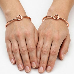 Può includere: Un paio di bracciali rigidi in metallo color oro rosa lucido, ciascuno caratterizzato da un distintivo meccanismo di chiusura a gancio. I bracciali presentano un design a fascia liscia e arrotondata che poggia comodamente attorno al polso.