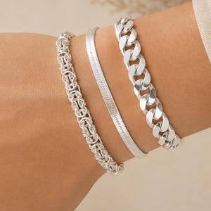 Peut inclure: Un ensemble de trois bracelets argentés portés ensemble. Le premier est une chaîne tressée complexe, le deuxième est une chaîne plate et lisse en forme d'arête de poisson, et le troisième est une grosse chaîne gourmette polie.