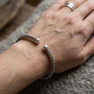 Può includere: Un bracciale a fascia color argento caratterizzato da una texture intrecciata simile a una corda lungo tutto il corpo. Il bracciale presenta un design aperto, rifinito con una sfera liscia e lucida su ciascuna delle due estremità.