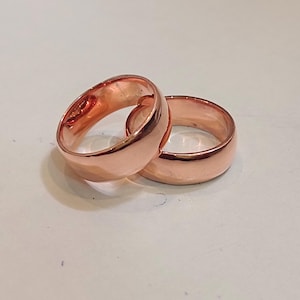 Könnte beinhalten: Zwei polierte Kupferringe mit einem glatten, abgerundeten Banddesign. Die Ringe haben eine warme, metallische Oberfläche mit einem reflektierenden Glanz. Beide Ringe sind schlicht, ohne Gravuren oder Steine, und liegen leicht übereinander.