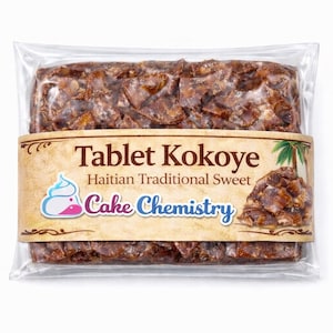 May include: A packaged block of Tablet Kokoye, a Haitian traditional sweet. The product is wrapped in clear plastic with a brown label featuring text and a logo for Cake Chemistry. The sweet is a dark brown color.