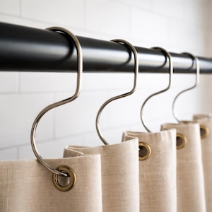 May include: A close-up of a shower curtain with metal hooks hanging from a black rod. The beige curtain has brass grommets. The hooks are silver and have a unique S-shape design.