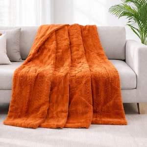 May include: An orange faux fur throw blanket draped over a gray sofa. The blanket has a textured pattern and is made of a soft, plush material. The sofa is in a living room setting with a plant in the background.