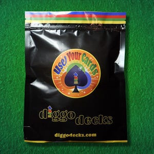 Puede incluir: Paquete negro con una parte superior a rayas colorida y un logotipo circular que dice "Use Your Cards". El logotipo presenta un diseño de pica. El texto "diggo decks" y "diggodecks.com" están impresos en el paquete.
