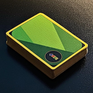 Puede incluir: Una baraja de cartas con un diseño geométrico verde en el reverso. Las cartas tienen un borde amarillo y un pequeño emblema circular con formas coloridas. La baraja está apilada sobre una superficie oscura.