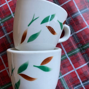 May include: Two white ceramic coffee mugs with a brown and green leaf design. The mugs are stacked on top of each other.