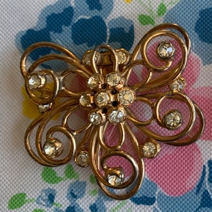 May include: A vintage gold-toned brooch with a floral design. The brooch features clear, sparkling rhinestones and intricate scrollwork. The brooch is set against a floral patterned background.