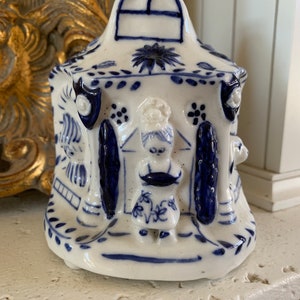 May include: A white ceramic figurine of a house with blue and white painted details. The house has a window and a small figure standing in front of it.