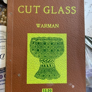 May include: A brown book with the title "AMERICAN CUT GLASS" by WARMAN. The cover features a green illustration of a cut glass goblet on a yellow background. The price of the book is $3.00.