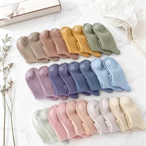 May include: A collection of colorful socks in various shades, including beige, pink, purple, blue, yellow, and green. Each pair of socks is neatly arranged, showcasing a ribbed texture and a small circular design on the top. The socks are presented on a white surface.